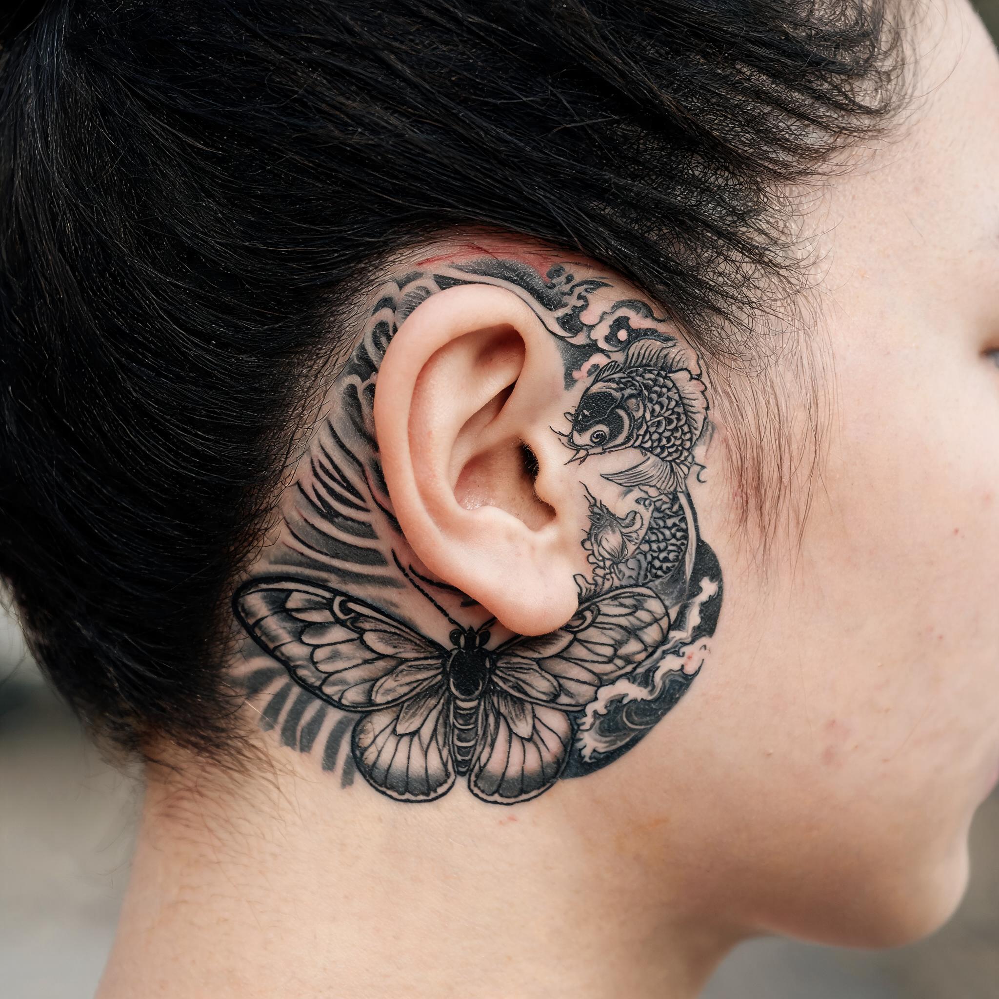 A black and grey Japanese irezumi death moth tattoo design with koi and waves, shown behind the ear on a real person.