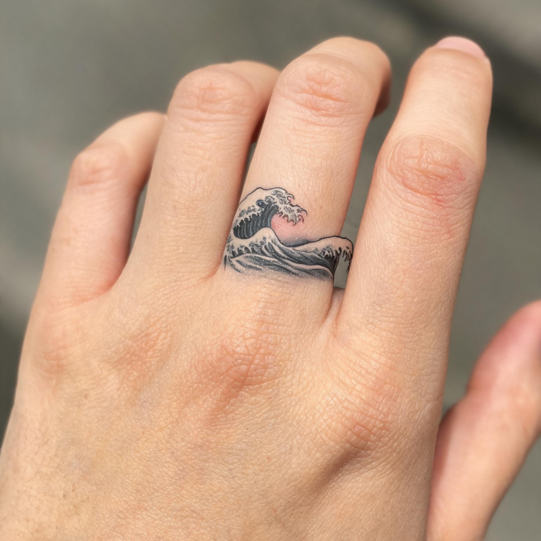 A close-up tattoo image of a Japanese wave design in elegant calligraphy script on a hand with fingers extended, shown on white.