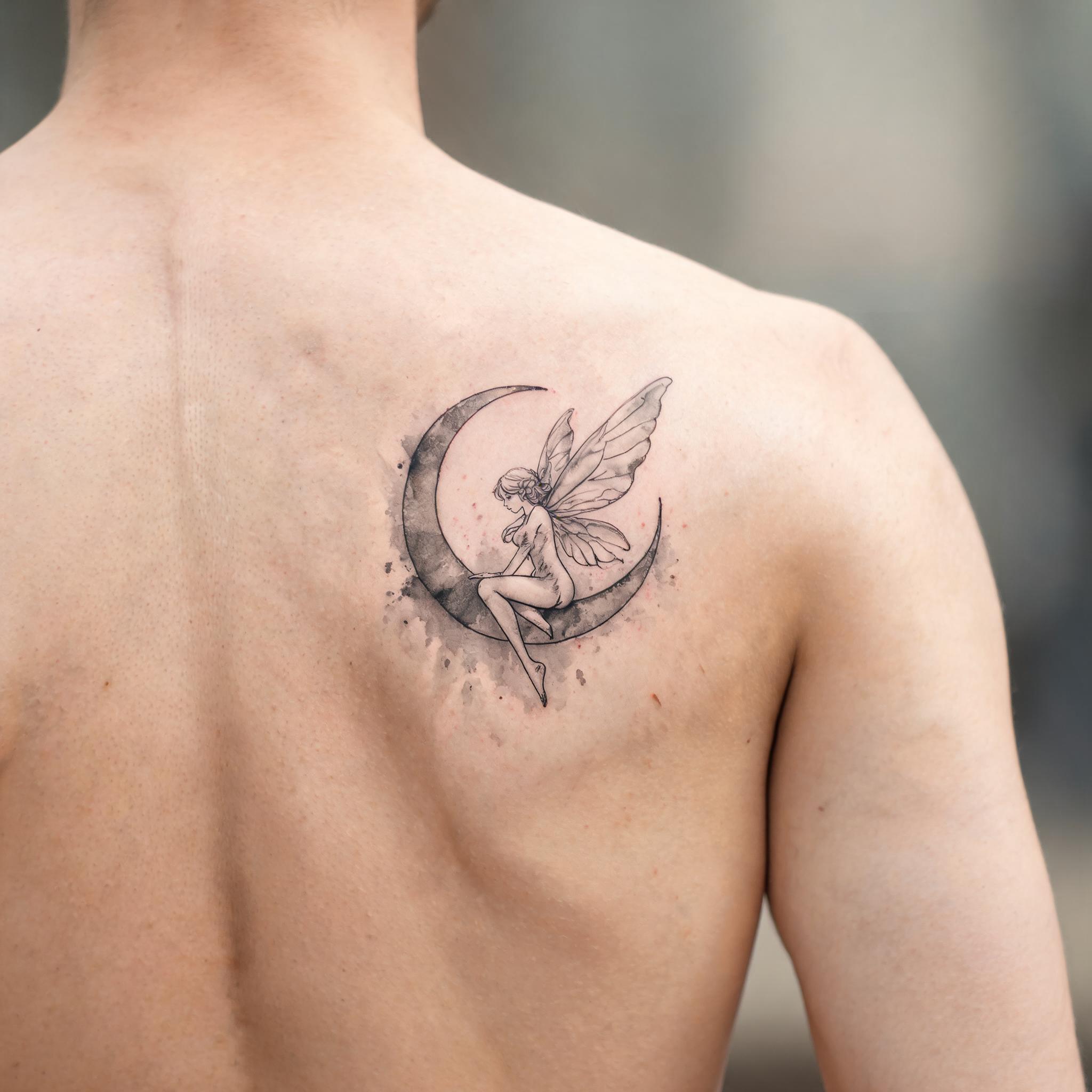 A black and grey watercolor tattoo image of a fairy sitting on a crescent moon, shown on a shoulder blade against neutral skin.
