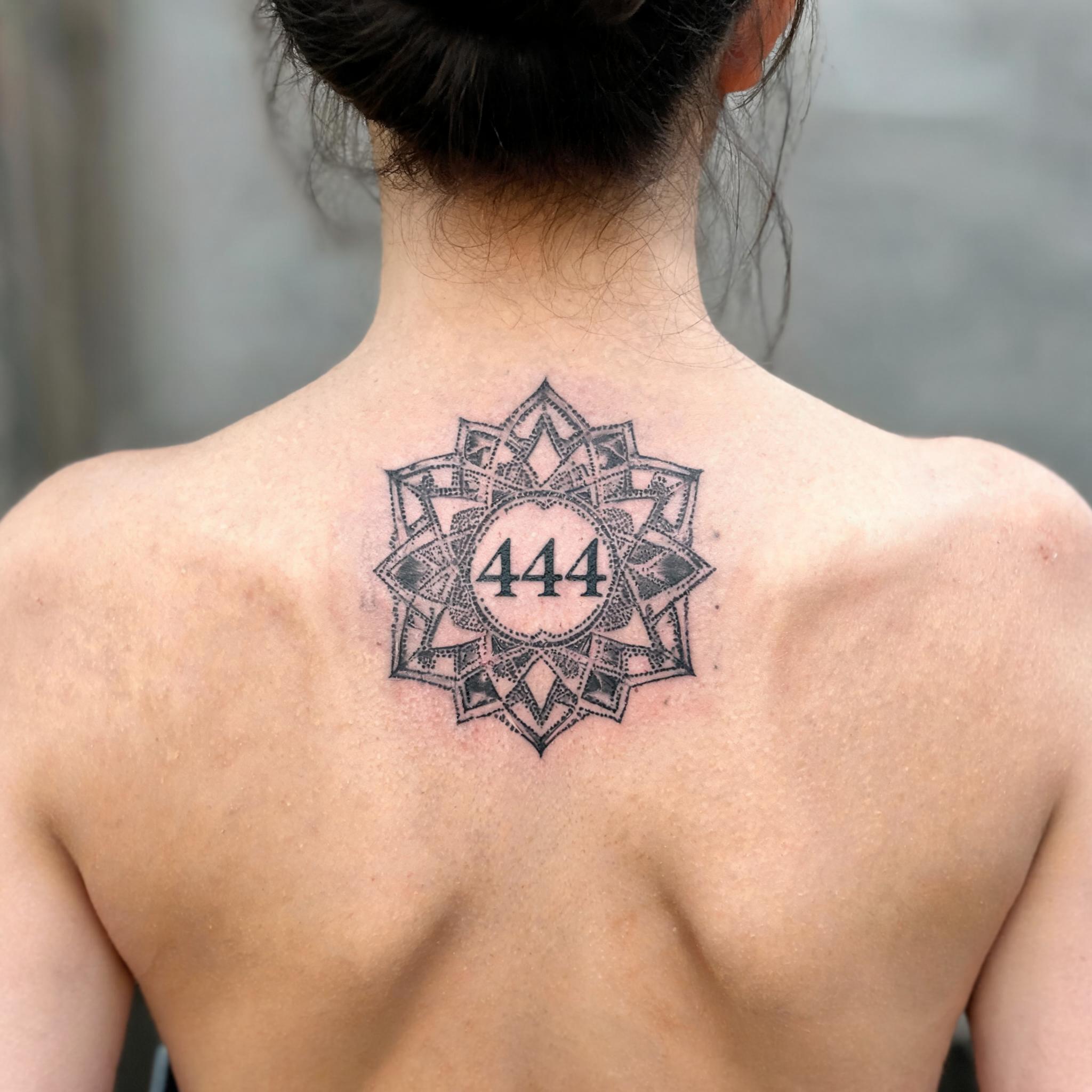 A geometric angel number tattoo image design with mandala symmetry and dotwork shading on a person's upper back, photographed on a plain background.