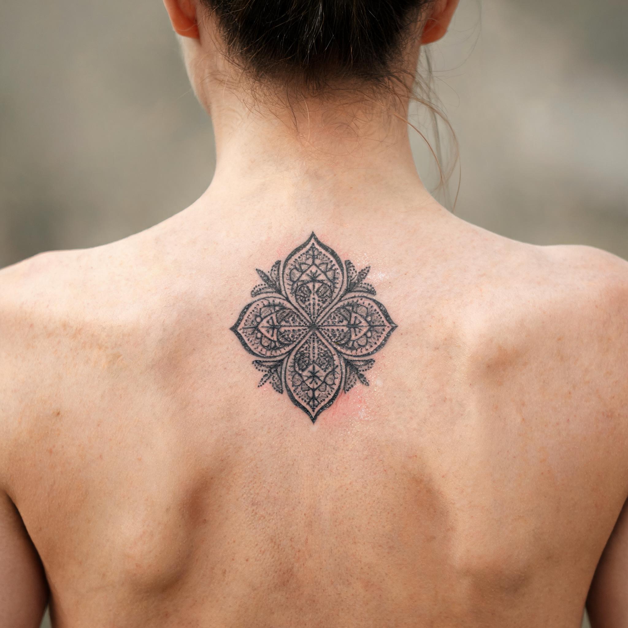 A geometric dotwork four leaf clover tattoo on the upper back, shown in black and grey on realistic skin.